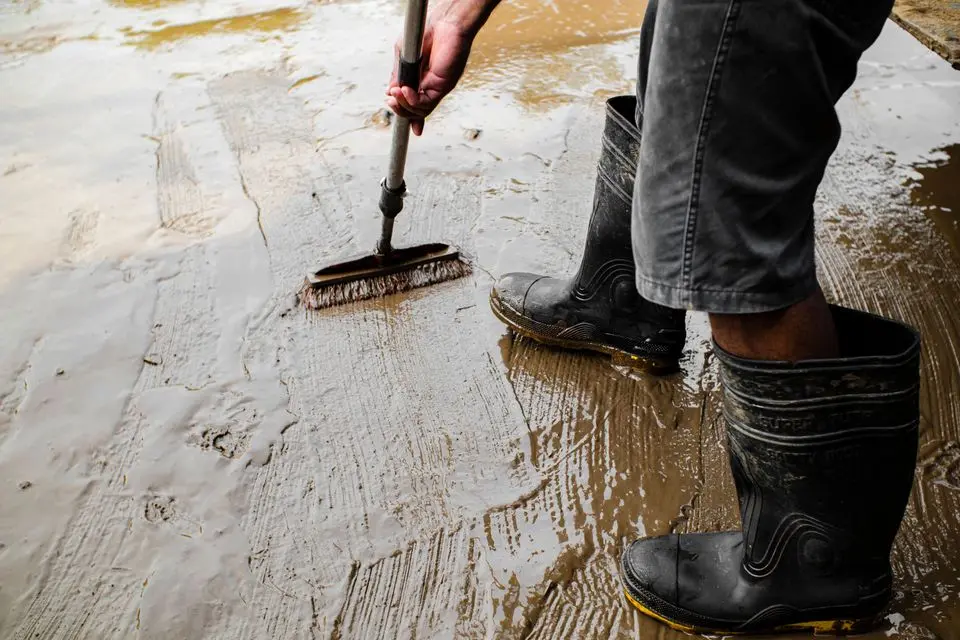 Worker in rubber boots scrubbing a wet concrete floor with a stiff brush