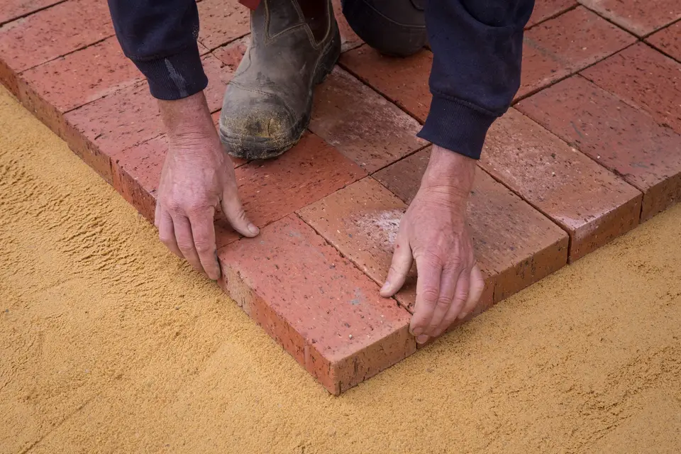 A worker in workwear laying brick pavers on a sand bed